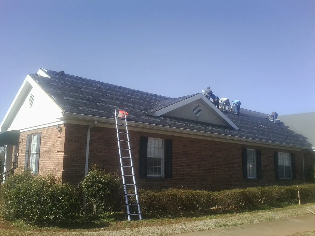 Crew tearing shingles off a brick ranch roof with a ladder set — Emergency Roof Repair work by our crew in Bryan